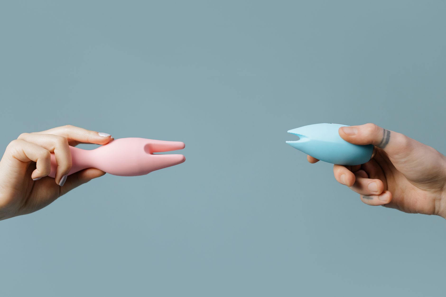 Two hands holding pink and blue silicone vibrators against a pastel background, symbolizing shared exploration and partnership.